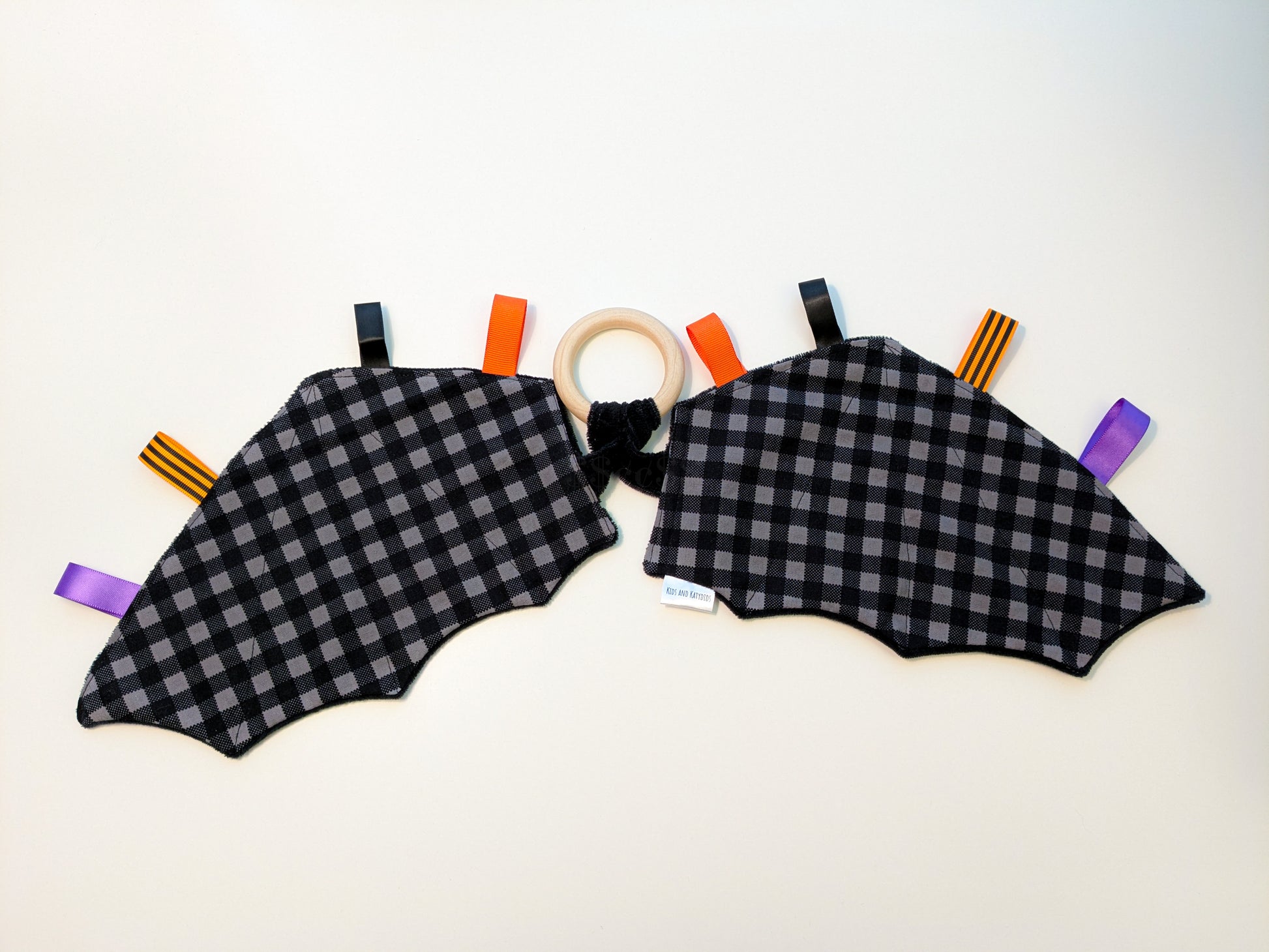 Black and gray checkered baby teether in the shape of bat-wings with colorful ribbon tags and a wooden ring on a white background 
