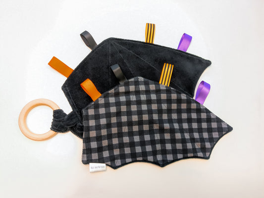 Black and gray checkered baby teether in the shape of bat-wings with colorful ribbon tags and a wooden ring on a white background 