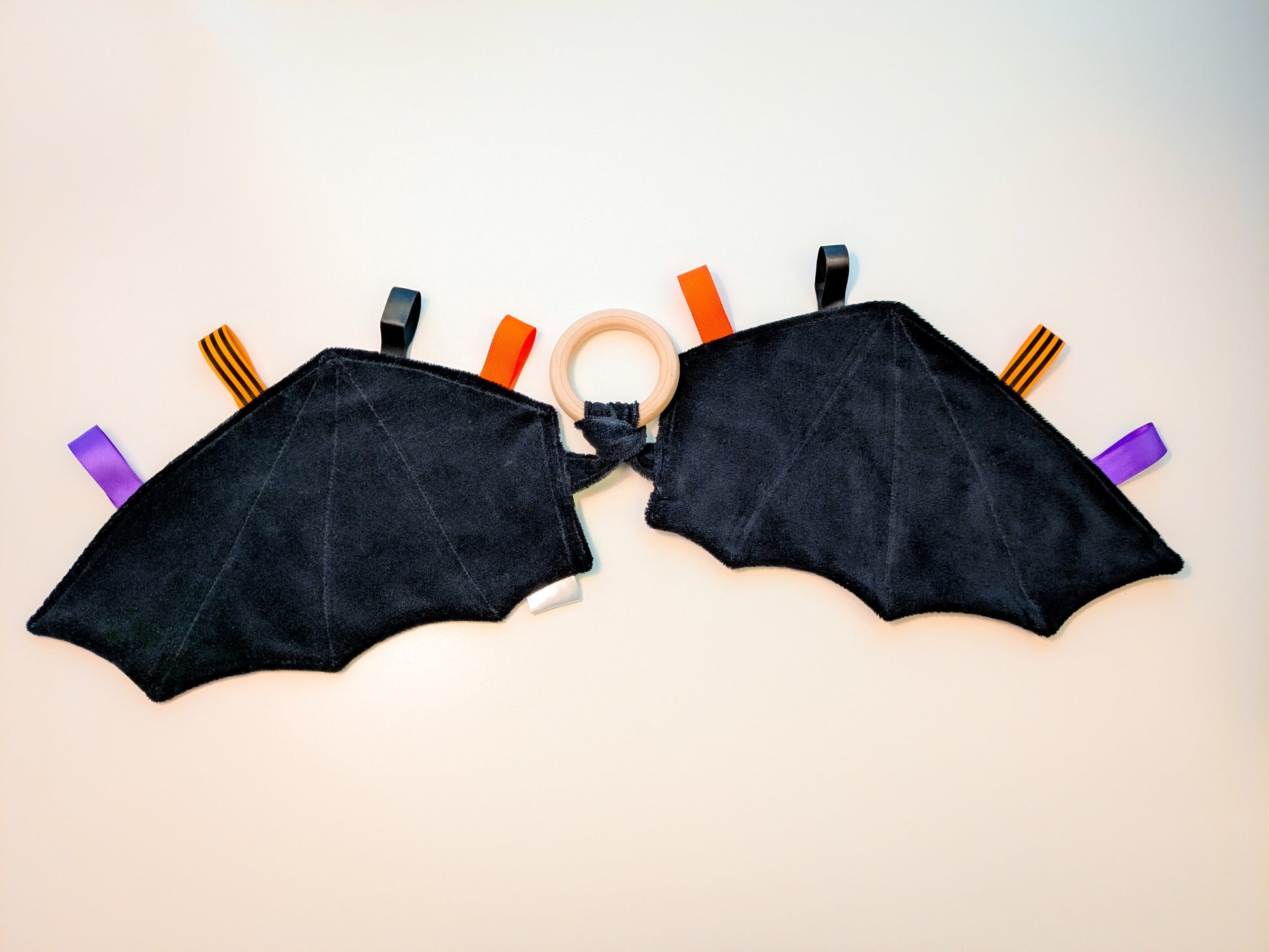 Black and gray checkered baby teether in the shape of bat-wings with colorful ribbon tags and a wooden ring on a white background 