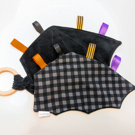 a video showing a black and gray checkered baby teether in the shape of bat-wings with colorful ribbon tags and a wooden ring on a white background 